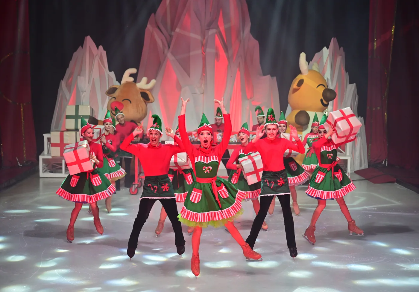 Skaters dressed as elves in red and green, with two giant reindeer costumes behind them on an ice set